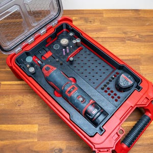 Peut inclure: Un outil rotatif sans fil Milwaukee M12 rouge et noir avec batterie, rangé dans une mallette de transport en plastique rouge avec un couvercle transparent. La mallette comporte un insert noir personnalisé avec des emplacements pour divers embouts.