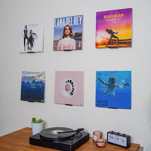 May include: Six square vinyl record album covers displayed on a wall in two rows of three. The covers feature artwork for Fleetwood Mac, Lana Del Rey, Bohemian Rhapsody, Kanye West, Mac Miller, and Nirvana. A black record player sits below.