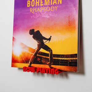 Puede incluir: Un soporte de exhibición de plástico rojo con el texto NOW PLAYING en relieve en la parte frontal, que sostiene una funda de disco de vinilo de Bohemian Rhapsody con la silueta de un cantante actuando en un escenario sobre un fondo de atardecer naranja y violeta.