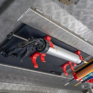 May include: A black and silver caulking gun secured to a black mounting board by two bright red plastic clips. The mounting board is attached to a diamond-plate metal surface. An additional set of empty red clips is visible below the gun.