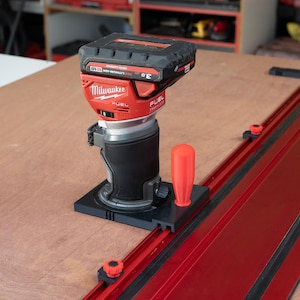 Puede incluir: Una fresadora compacta inalámbrica Milwaukee M18 Fuel roja y negra con batería de 3.0 Ah, montada sobre una base negra con mango naranja, colocada sobre un riel guía de metal rojo largo en una superficie de madera plana.