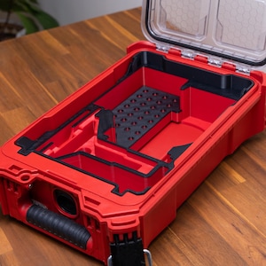 May include: A rectangular red storage case with an open lid and a black interior organizer tray. The tray features multiple compartments, including a section with a grid of small circular holes. A black rubberized handle is attached to the front.