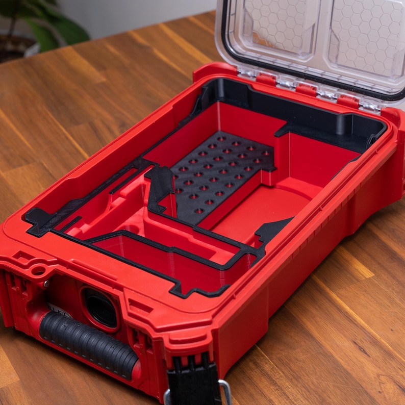May include: A rectangular red storage case with an open lid and a black interior organizer tray. The tray features multiple compartments, including a section with a grid of small circular holes. A black rubberized handle is attached to the front.