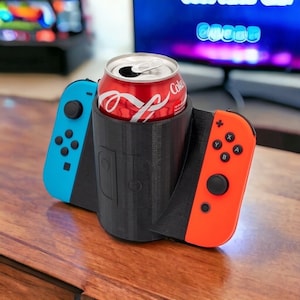 May include: A black 3D printed holder for a can of soda, with a Nintendo Switch Joy-Con on each side. The holder is designed to fit a standard 12-ounce can. The Joy-Cons are in the colors blue and red.
