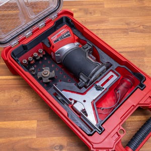 Op de afbeelding: Een rode Milwaukee M18 FTR draadloze compacte bovenfrees in een bijpassende rode harde kunststof koffer. De koffer bevat schuimrubberen inzetstukken voor de frees, een metalen zijgeleider, een doorzichtige stofkap en diverse freesbits.