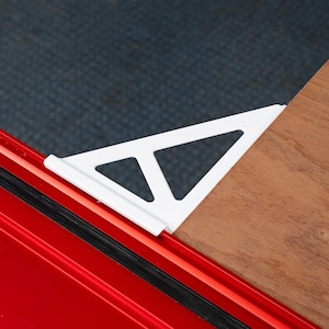 May include: A white triangular support bracket with a hollow geometric cutout design. The bracket is positioned to secure a light brown wooden board against a bright red metal frame, providing structural stability for the connected components.