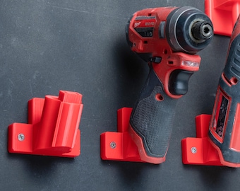 Könnte beinhalten: Drei leuchtend rote Wandhalterungen aus Kunststoff, befestigt mit silbernen Schrauben. Eine Halterung ist leer, während die anderen beiden rote und schwarze Akku-Elektrowerkzeuge tragen, darunter ein Milwaukee M12 Schlagschrauber.