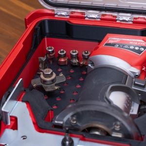 May include: A red Milwaukee M18 FTR cordless compact router stored inside a red and black plastic carrying case. The case features a black insert with multiple circular cutouts holding various metal router bits and cutting accessories.