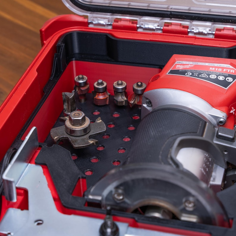 May include: A red Milwaukee M18 FTR cordless compact router stored inside a red and black plastic carrying case. The case features a black insert with multiple circular cutouts holding various metal router bits and cutting accessories.