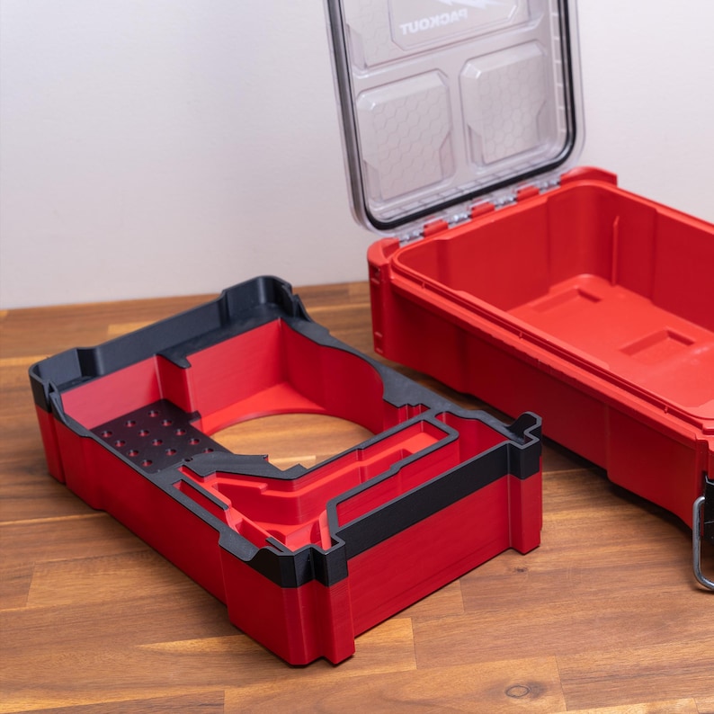 May include: A red and black plastic tool box insert with custom molded compartments and a grid of small holes, positioned next to an open red tool box with a clear lid featuring the word PACKOUT printed in white on the interior surface.