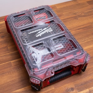 May include: A red Milwaukee Packout organizer with a clear, hexagonal-patterned lid. The rectangular storage case features a black handle and a transparent top that reveals internal compartments, with the Milwaukee Packout logo printed in white.