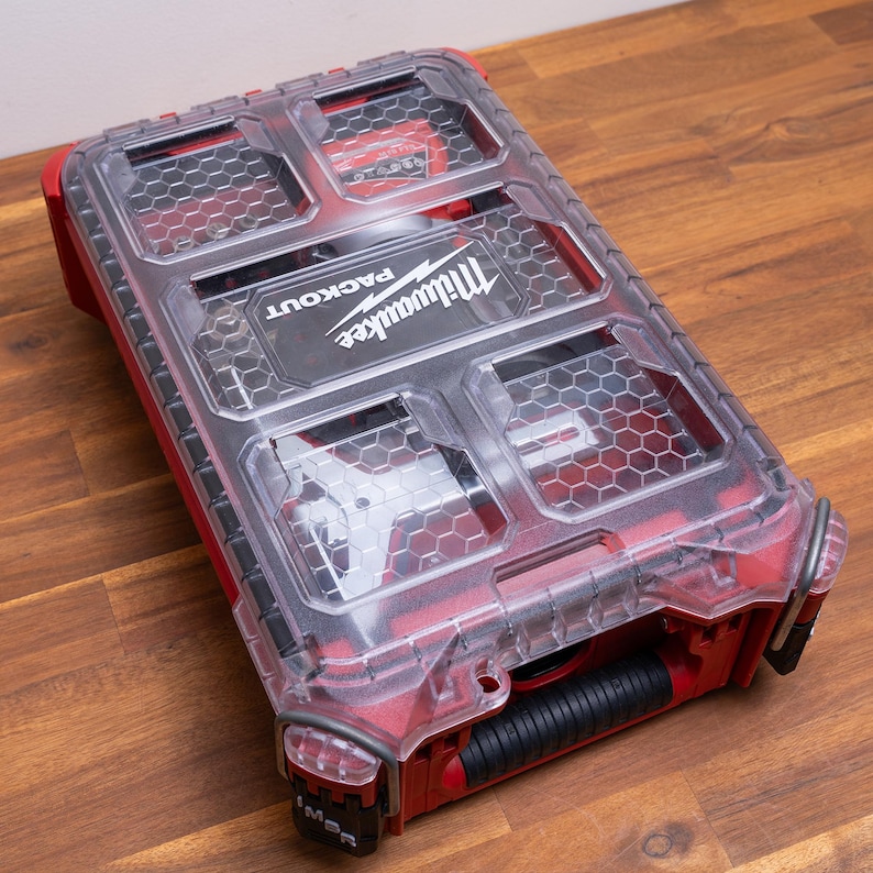 May include: A red Milwaukee Packout organizer with a clear, hexagonal-patterned lid. The rectangular storage case features a black handle and a transparent top that reveals internal compartments, with the Milwaukee Packout logo printed in white.