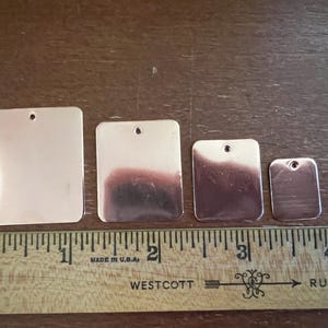 May include: Four blank, rectangular metal tags in varying sizes, each with a small hole at the top. The tags are arranged on a wooden surface next to a ruler marked in inches. The metal has a shiny, reflective surface.