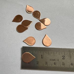 May include: Copper-colored teardrop-shaped metal charms scattered on a light gray surface. A metal ruler with inch markings is placed near the charms. The charms are approximately 1 inch in length.