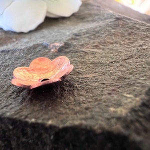 May include: A small, copper-colored flower-shaped charm with five petals and a hole in the center. The charm is lying on a rough, gray surface.