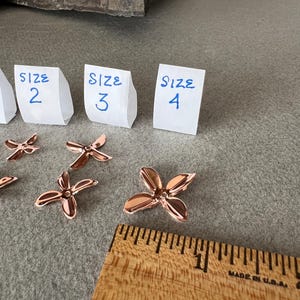 May include: Rose gold-colored, four-petal flower-shaped components. Three paper tags labeled "SIZE 2", "SIZE 3", and "SIZE 4" are visible. A wooden ruler with inch measurements is also present, indicating the size of the components.