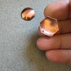 May include: Two copper-coloured metal hexagon shapes with a hole in the centre. The hexagon on the left is slightly rounded. The hexagon on the right is flat.