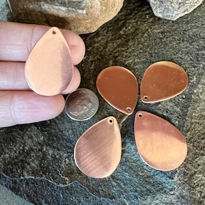 May include: Five copper teardrop-shaped metal blanks with a hole at the top. The blanks are arranged on a dark grey stone surface.