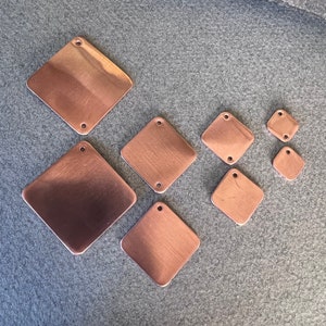 May include: Assortment of seven copper-colored metal diamond-shaped tags with two holes in each. The tags are arranged in a staggered pattern on a gray surface.