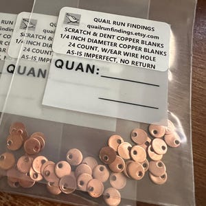 May include: Copper blanks with ear wire holes are displayed in clear plastic bags. The product label reads "SCRATCH & DENT COPPER BLANKS 1/4 INCH DIAMETER COPPER BLANKS 24 COUNT, W/EAR WIRE HOLE AS-IS IMPERFECT, NO RETURN."
