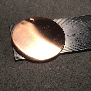 May include: A round, copper-colored metal disc with a small hole in the center. The disc is about 4 inches in diameter and is lying on a ruler.