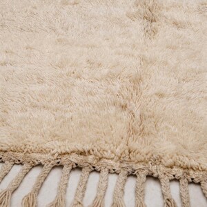 May include: A thick, cream-colored rug with a high-pile, shaggy texture. The rug features a woven base and a decorative edge finished with long, braided fringe tassels that hang from the bottom border of the textile piece.