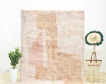 May include: A large, rectangular rug with a cream-colored background and abstract geometric shapes in shades of beige and dusty rose. The rug has a shaggy texture and a fringe along the bottom edge. A small plant in a white pot is on the left, and a straw hat and bag are on the right.