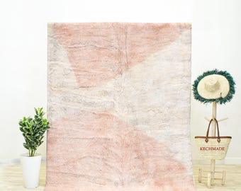 May include: A large, rectangular rug with a geometric design in shades of pink and off-white. The rug has a textured appearance and a fringe along the bottom edge. A small plant in a white pot is on the left.