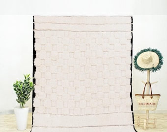 May include: A white Moroccan rug with a black border and a woven pattern of squares. The rug has a fringe border.