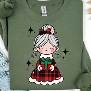 Può includere: Una felpa verde oliva con un'illustrazione a cartoni animati di una ragazza con i capelli grigi, un fiocco rosso e un vestito a quadri rosso e nero. Tiene in mano una confezione regalo verde con un fiocco rosso. Accenti di stelle nere circondano l'illustrazione.