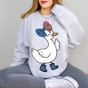 Peut inclure: Un sweat-shirt gris avec un canard blanc portant un n&oelig;ud rouge, blanc et bleu et des bottes de pluie bleues. Le canard sourit et a un bec paillet&eacute; d'or.