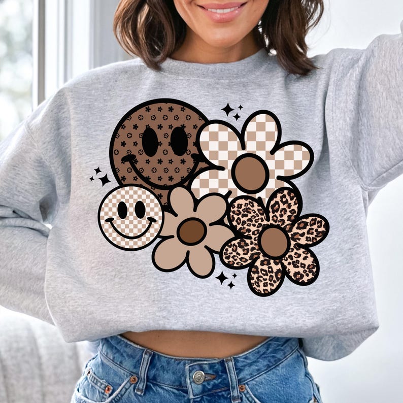 Peut inclure: Un sweat-shirt gris avec un motif graphique brun, noir et blanc repr&eacute;sentant un visage souriant et des fleurs. Les fleurs ont des motifs diff&eacute;rents, notamment &agrave; carreaux, &agrave; imprim&eacute; l&eacute;opard et &agrave; fleurs.