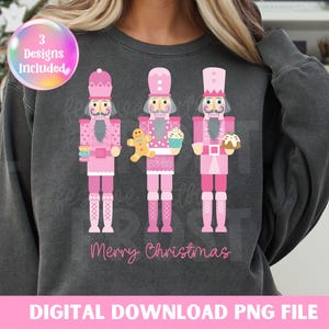 May include: A gray sweatshirt with a pink, white, and gray design of three nutcracker figures holding Christmas treats. The text "Merry Christmas" is printed below the figures.