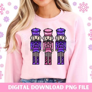 May include: A pink sweatshirt featuring a graphic of three nutcracker figures. The nutcrackers are wearing purple, pink, and blue hats and outfits. The graphic is centred on the sweatshirt.