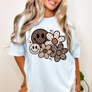 Peut inclure: Un t-shirt bleu clair avec un motif graphique brun et noir repr&eacute;sentant un visage souriant et des fleurs. Les fleurs ont des motifs diff&eacute;rents, notamment &agrave; carreaux, &agrave; imprim&eacute; l&eacute;opard et &agrave; motif floral brun.