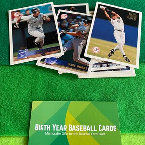 May include: A collection of baseball cards featuring various players, including Don Mattingly and Wade Boggs. The cards are from the 1990s and feature a blue and white color scheme. The cards are arranged in a stack, with the top card showing a player in a batting stance. The cards are on a green surface. The text "BIRTH YEAR BASEBALL CARDS" and "Memorable Gifts for the Baseball Enthusiast" is visible on a green and yellow card.