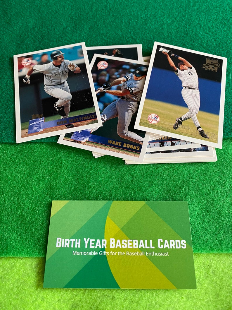 May include: A collection of baseball cards featuring various players, including Don Mattingly and Wade Boggs. The cards are from the 1990s and feature a blue and white color scheme. The cards are arranged in a stack, with the top card showing a player in a batting stance. The cards are on a green surface. The text "BIRTH YEAR BASEBALL CARDS" and "Memorable Gifts for the Baseball Enthusiast" is visible on a green and yellow card.