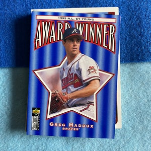 May include: A baseball card featuring Greg Maddux, a baseball player for the Atlanta Braves. The card is blue and red with a white star and the text "1995 N.L. Cy Young Award Winner".