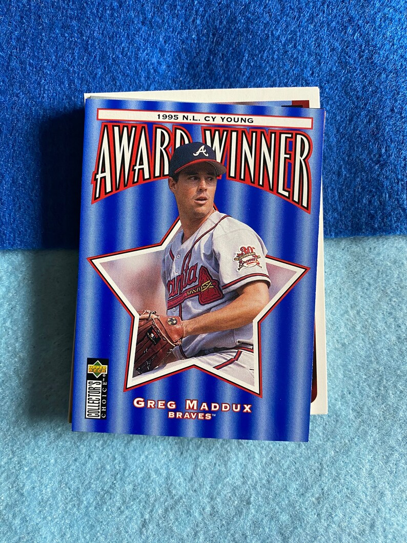 May include: A baseball card featuring Greg Maddux, a baseball player for the Atlanta Braves. The card is blue and red with a white star and the text "1995 N.L. Cy Young Award Winner".