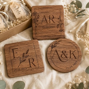Puede incluir: Posavasos de madera con iniciales y fechas grabadas. Un posavasos cuadrado presenta "AR" y "12/10/2023", otro "ER" y "12.10.2023", y uno redondo con "A&K" y "12.08.2023". Los posavasos están rodeados de elementos decorativos y bolsas de regalo.