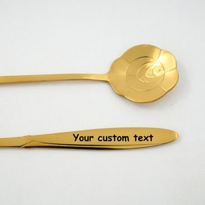 May include: Two gold-colored metal spoons. One spoon has a rose-shaped bowl. The other spoon has the text "Your custom text" engraved on the handle.