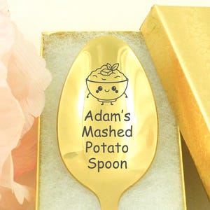 May include: A gold-colored spoon engraved with "Adam's Mashed Potato Spoon" and a cartoon image of mashed potatoes. The spoon is in a gold gift box with a light green interior, next to a pink flower.