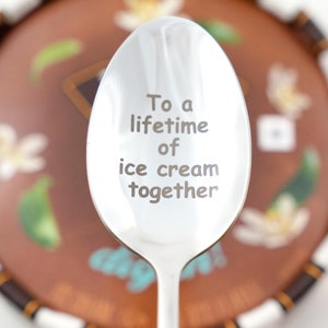 Puede incluir: Una cuchara de plata con la inscripci&oacute;n "To a lifetime of ice cream together" grabada en ella. La cuchara est&aacute; enfocada, y un plato de postre marr&oacute;n y blanco est&aacute; desenfocado en el fondo.