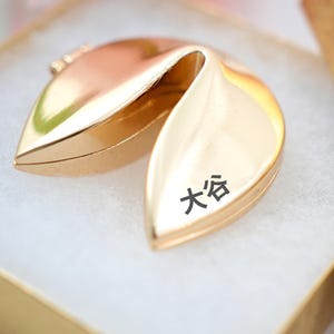 May include: A gold-colored fortune cookie with the Japanese characters 大谷 printed on the front, resting inside a gold box with a white interior. The cookie is open, revealing its interior. The box is also gold.