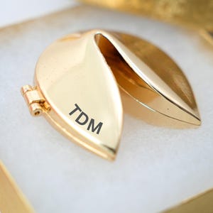 May include: A gold-colored fortune cookie-shaped locket, partially open, revealing the interior. The locket is in a gold box with a white interior. The initials "TDM" are engraved on the front of the locket.