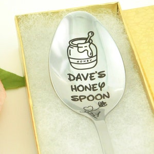 May include: A silver spoon with a honey jar and bee design engraved on it. The spoon is in a white box with a gold trim. The text on the spoon reads "Dave's Honey Spoon".