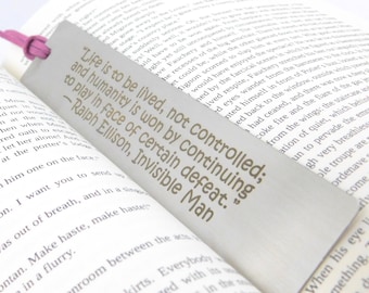 May include: A silver bookmark with a pink ribbon. The bookmark features a quote by Ralph Ellison: "Life is to be lived, not controlled; and humanity is won by continuing to play in face of certain defeat." - Ralph Ellison, Invisible Man