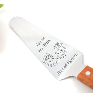 Puede incluir: Servidor de pastel de acero inoxidable con mango de madera. El servidor presenta el texto "You're my little" y "Slice of Heaven" junto con dos rebanadas de pastel de dibujos animados tomadas de la mano.