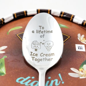 Puede incluir: Una cuchara de plata con las palabras "To a lifetime of Ice Cream Together" grabadas. La cuchara tiene un cono de helado de dibujos animados y una paleta con caras.