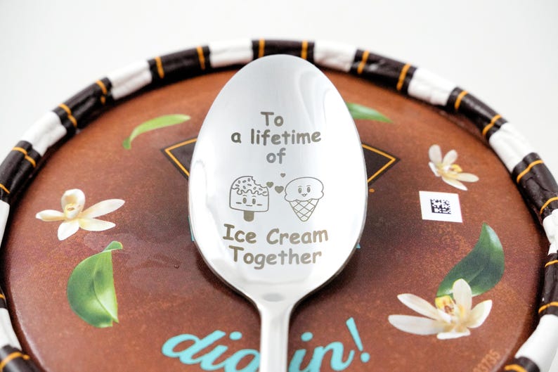 Puede incluir: Una cuchara de plata con las palabras "To a lifetime of Ice Cream Together" grabadas. La cuchara tiene un cono de helado de dibujos animados y una paleta con caras.